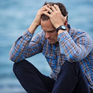 A man in a plaid shirt sits by the water looking distressed, symbolizing stress.